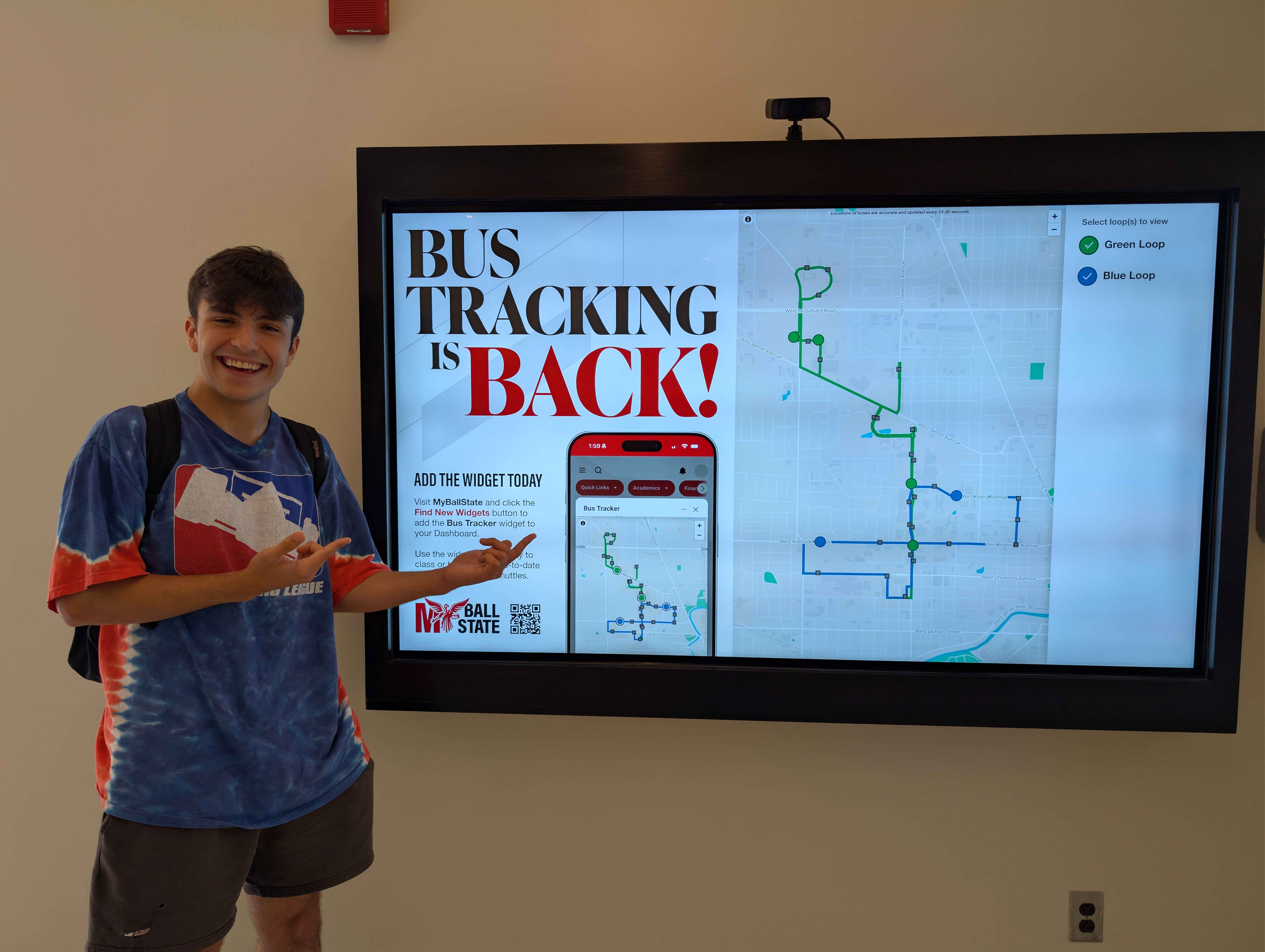 Lucas standing in front of a TV that is displaying a live advert of the Ball State Bus Tracker widget when it was first released to students in the beginning of the fall semester.